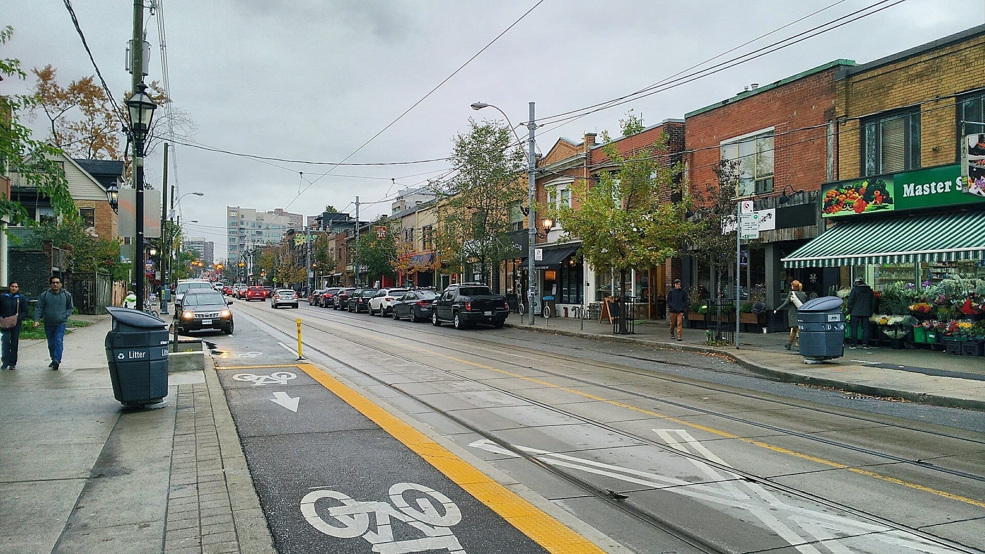 วิวถนนในย่าน Roncesvalles Village เมือง Toronto ในวันที่มีเมฆมาก แสดงให้เห็นร้านค้าอิสระ รถยนต์จอดอยู่ เลนจักรยาน และคนเดินถนน