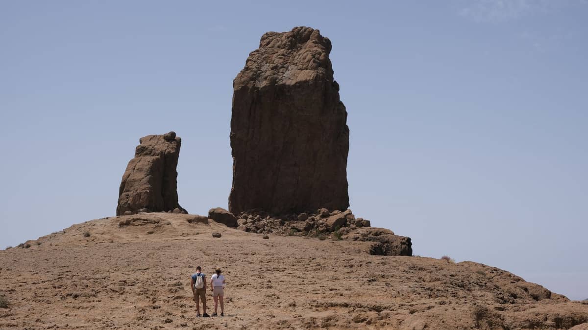 Трое туристов стоят на плато перед впечатляющим вулканическим монолитом Roque Nublo на фоне ясного голубого неба Гран-Канарии.