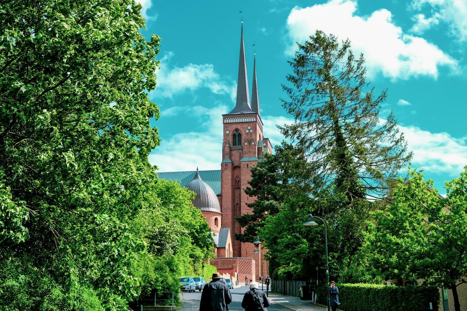 Le due torri gemelle della Cattedrale di Roskilde si innalzano sopra lussureggianti alberi verdi sotto un cielo azzurro brillante, con persone che camminano verso la storica chiesa in mattoni.