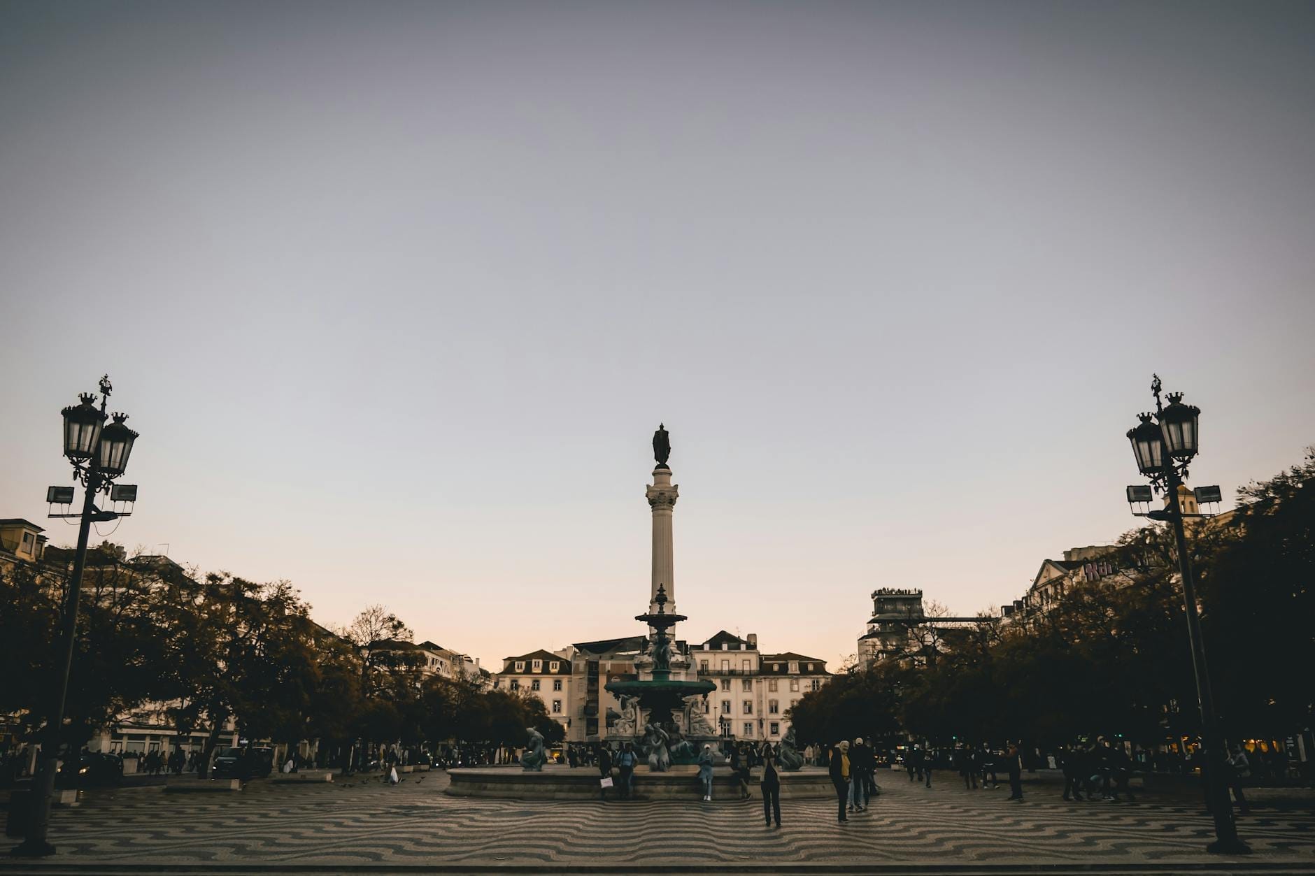 Vista panoramica della Piazza del Rossio al tramonto con la colonna centrale, il lastricato a onde, gli edifici storici, i lampioni e la gente raccolta a Lisbona, Portogallo.