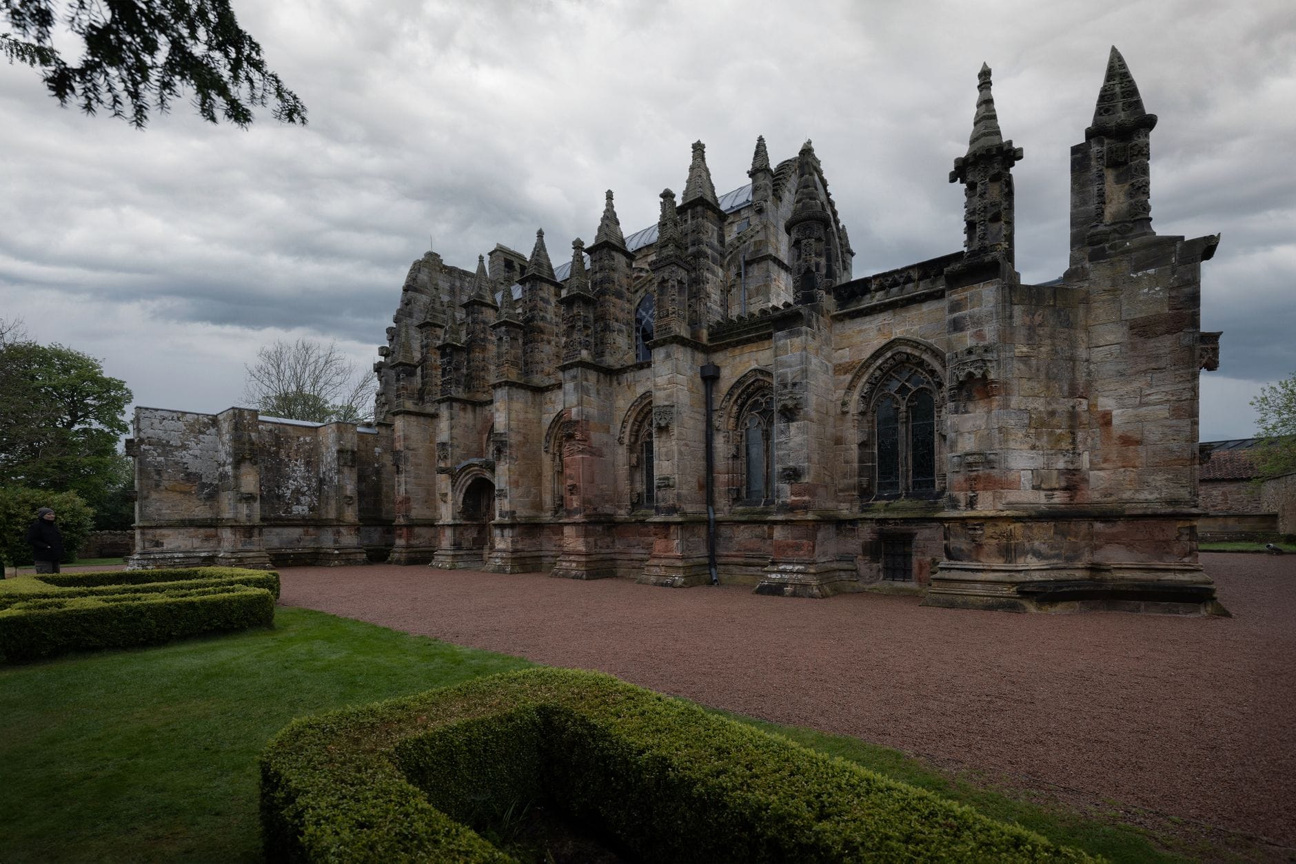 Weitwinkelfoto der Außenansicht der Rosslyn Chapel mit charakteristischen Steinmetzarbeiten, Türmen, gotischen Fenstern und kunstvollen Schnitzereien, aufgenommen von den Gartenanlagen unter dramatischem Himmel.