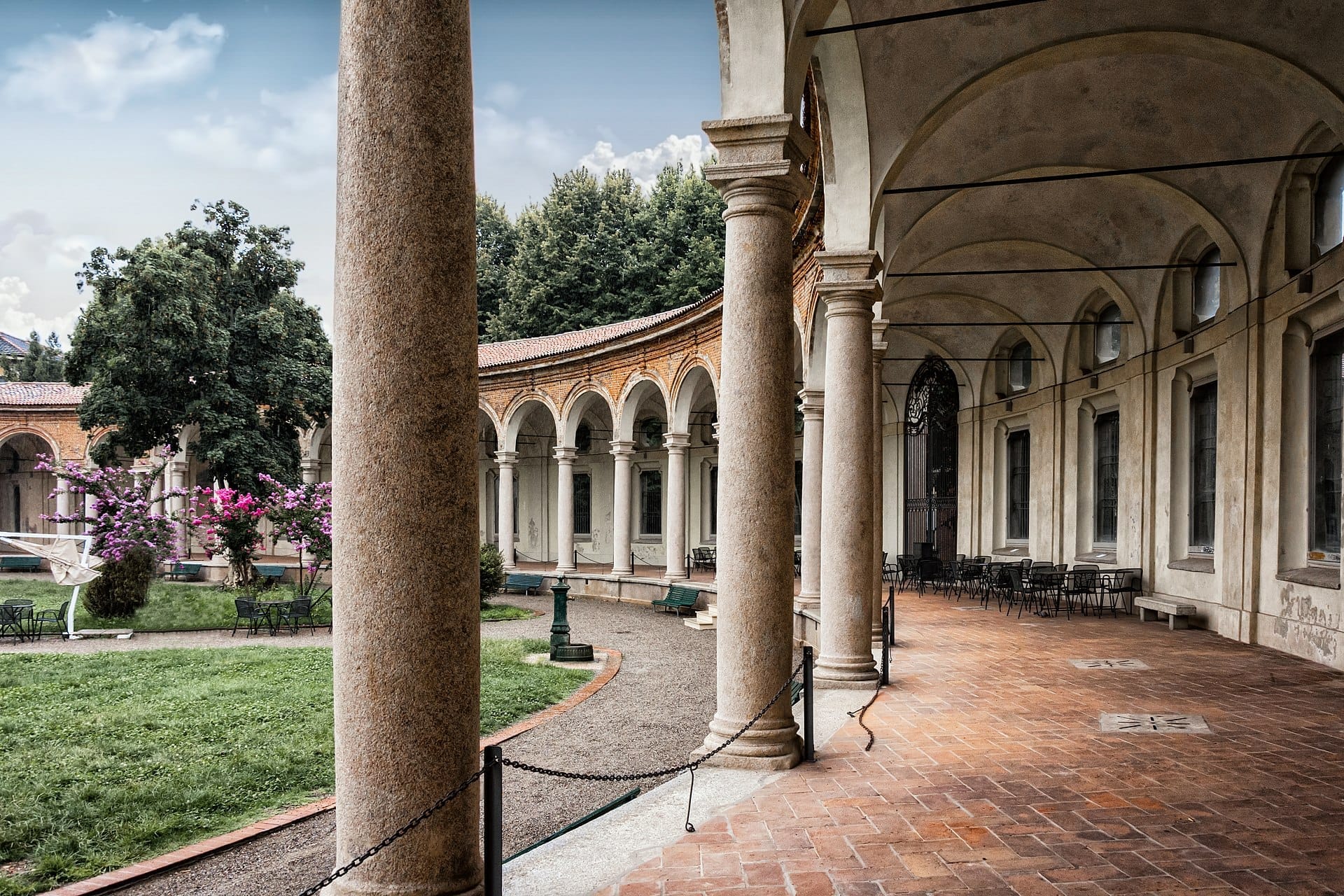 Vista del colonnato ad archi della Rotonda della Besana e del tranquillo giardino centrale con fiori vivaci e sedie vuote all'aperto in una giornata nuvolosa.