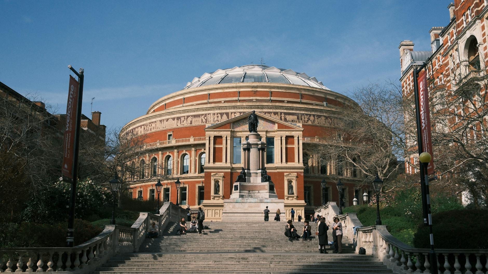 Vista panoramica della Royal Albert Hall a Londra: l'iconica facciata circolare in mattoni rossi, la statua centrale e la grande scalinata in una giornata di sole.
