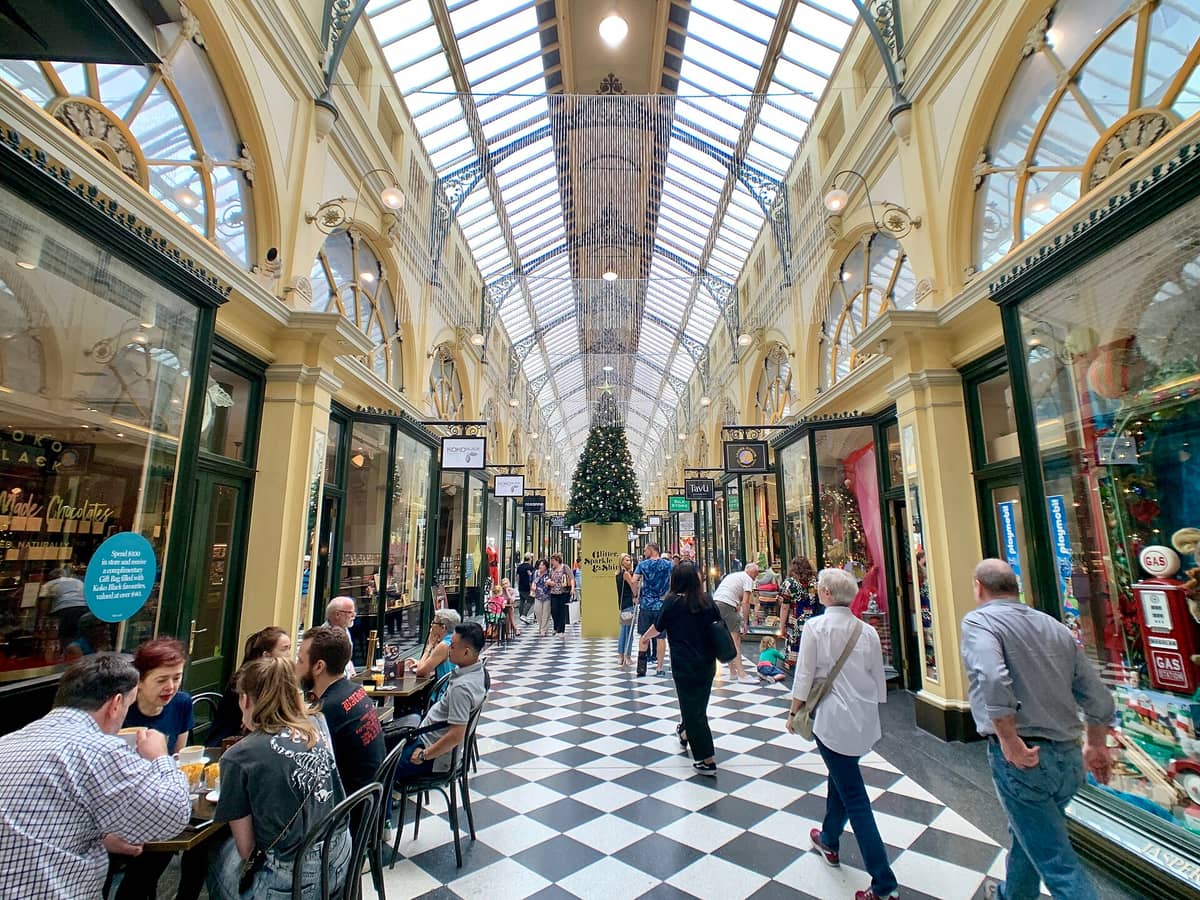 Vista interior de la Royal Arcade de Melbourne con su ornamentada arquitectura victoriana, techo de vidrio, piso en damero, fachadas de tiendas y personas paseando y comiendo dentro de la histórica galería.