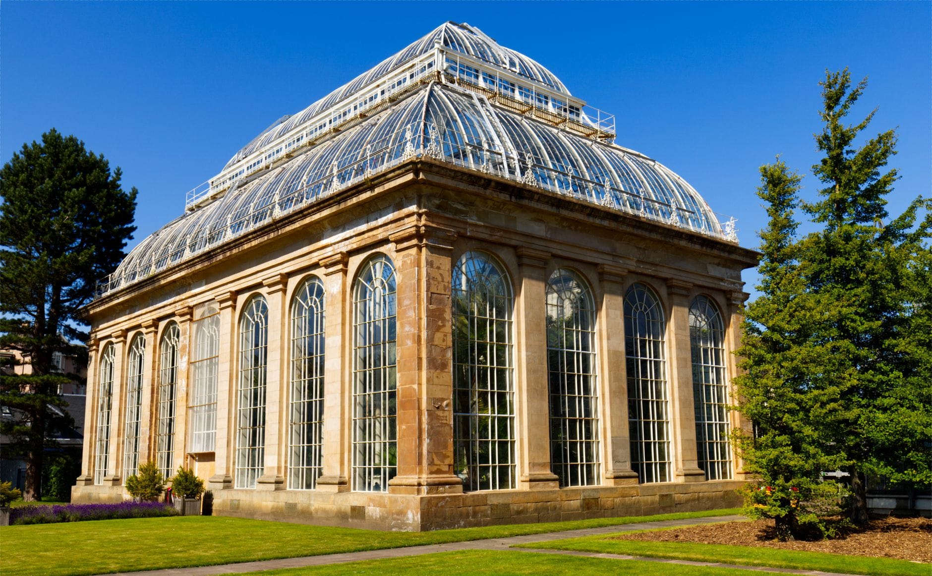Blick auf das viktorianische Palmenhaus im Royal Botanic Garden Edinburgh mit blauem Himmel, grünen Bäumen und Rasenfläche.