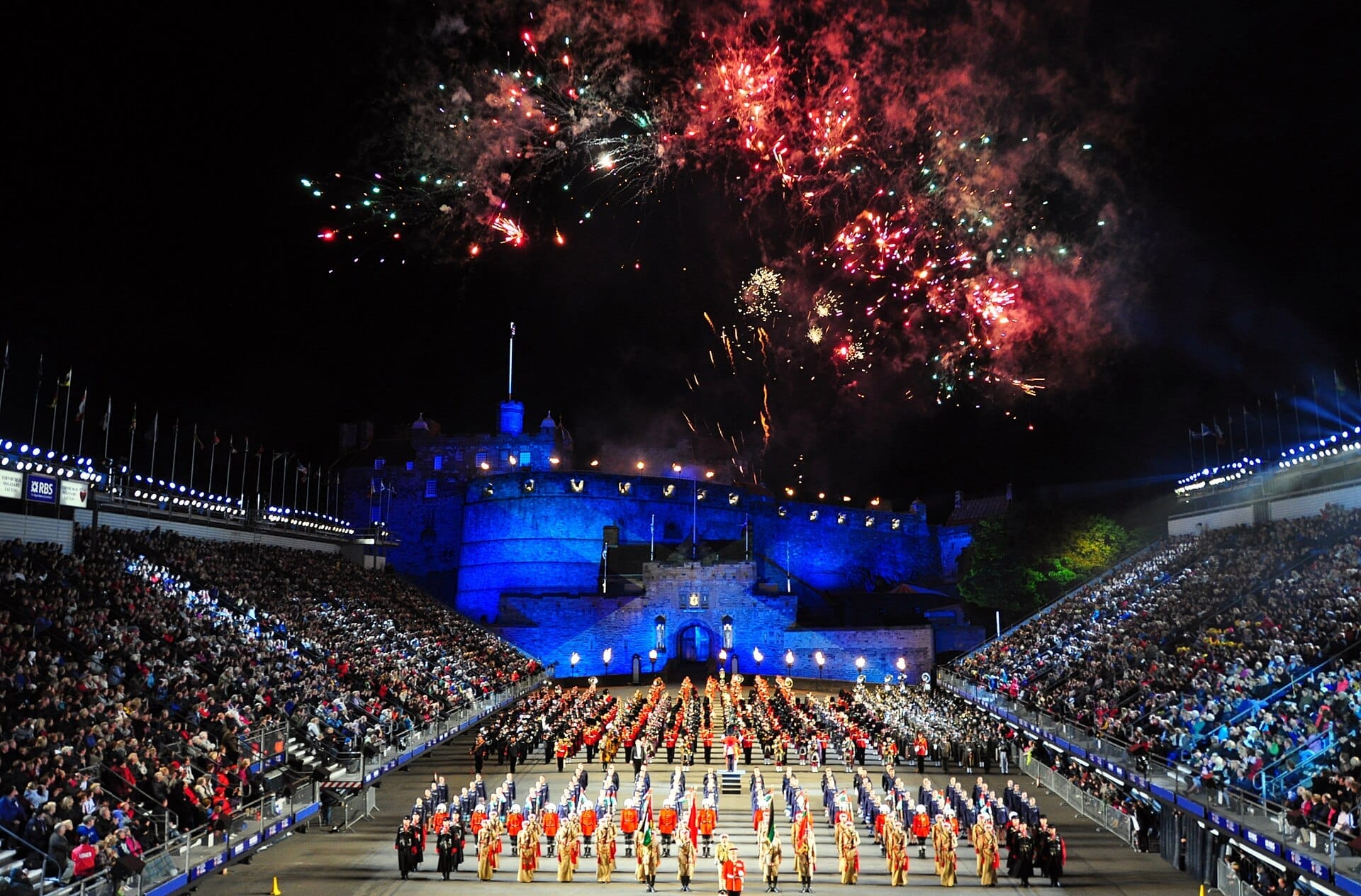 Das Royal Edinburgh Military Tattoo zeigt Musikkapellen in bunten Uniformen auf der Esplanade, während das Edinburgh Castle beleuchtet ist und Feuerwerk den Nachthimmel erhellt.