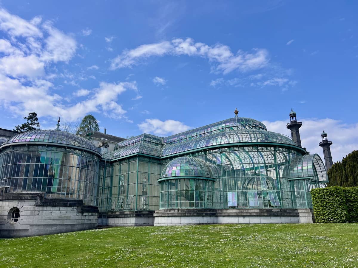 Vista panorâmica das Estufas Reais de Laeken mostrando a ornamentada estrutura de vidro e ferro sob um céu azul, rodeada por gramado verde.