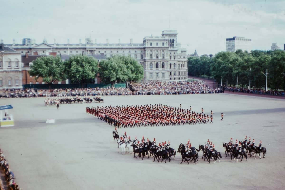 Um grande desfile real com soldados e guardas a cavalo em formação em Horse Guards Parade, Londres, assistido por grandes multidões e edifícios históricos.