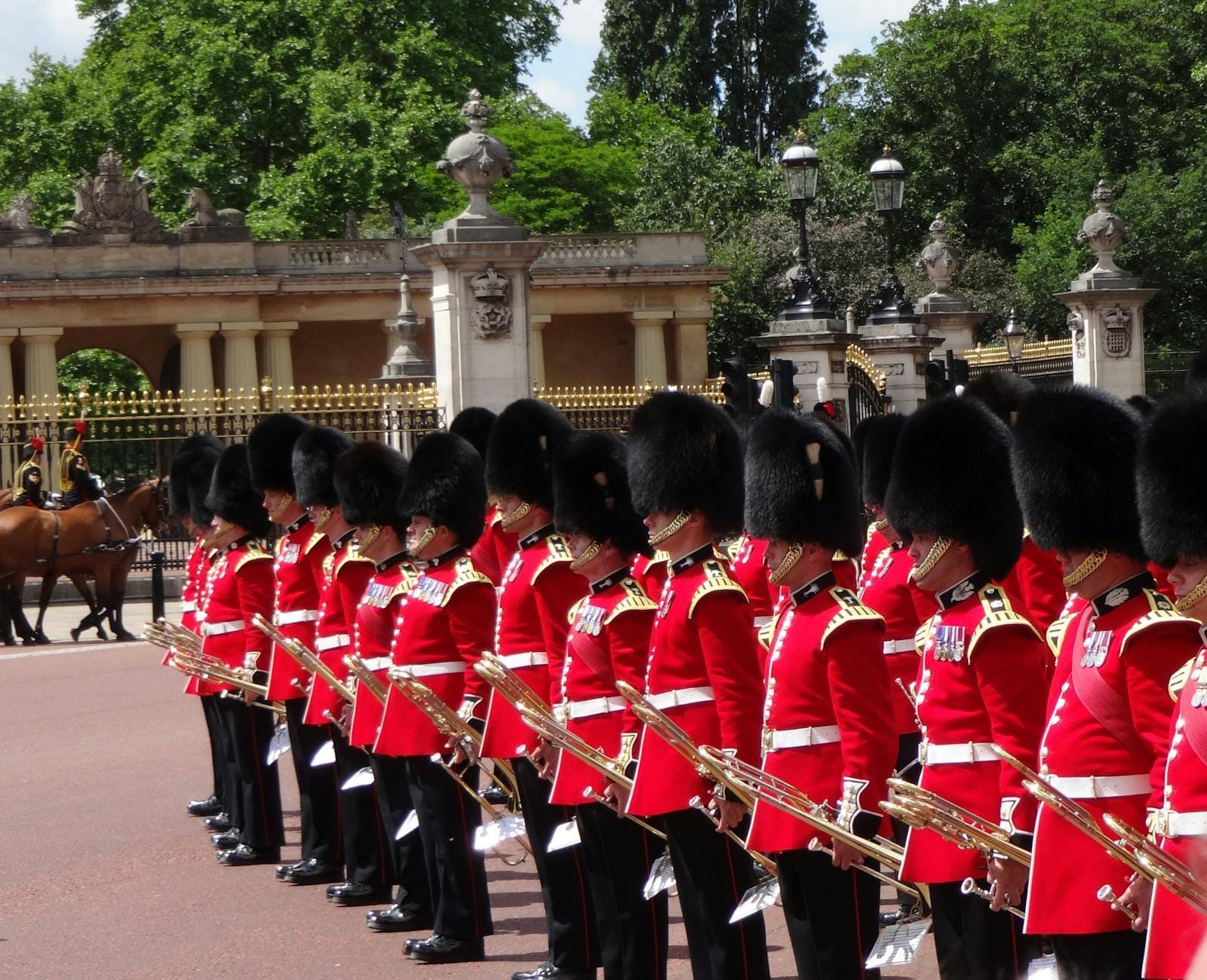 Uma fila de guardas reais em uniformes vermelhos e chapéus de pele de urso formados do lado de fora do Palácio de Buckingham, com portões ornamentados e vegetação ao fundo.
