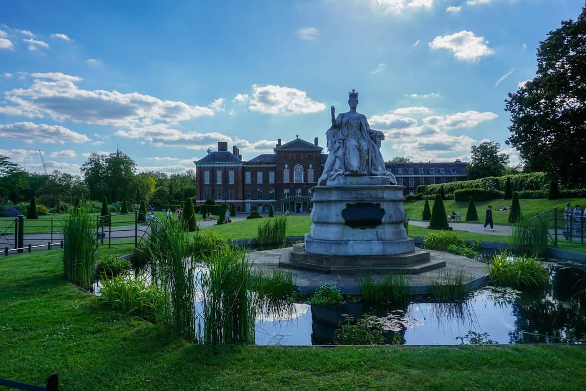 Estátua da Rainha Vitória em frente ao Kensington Palace com jardins formais e um lago refletivo sob um céu ensolarado.