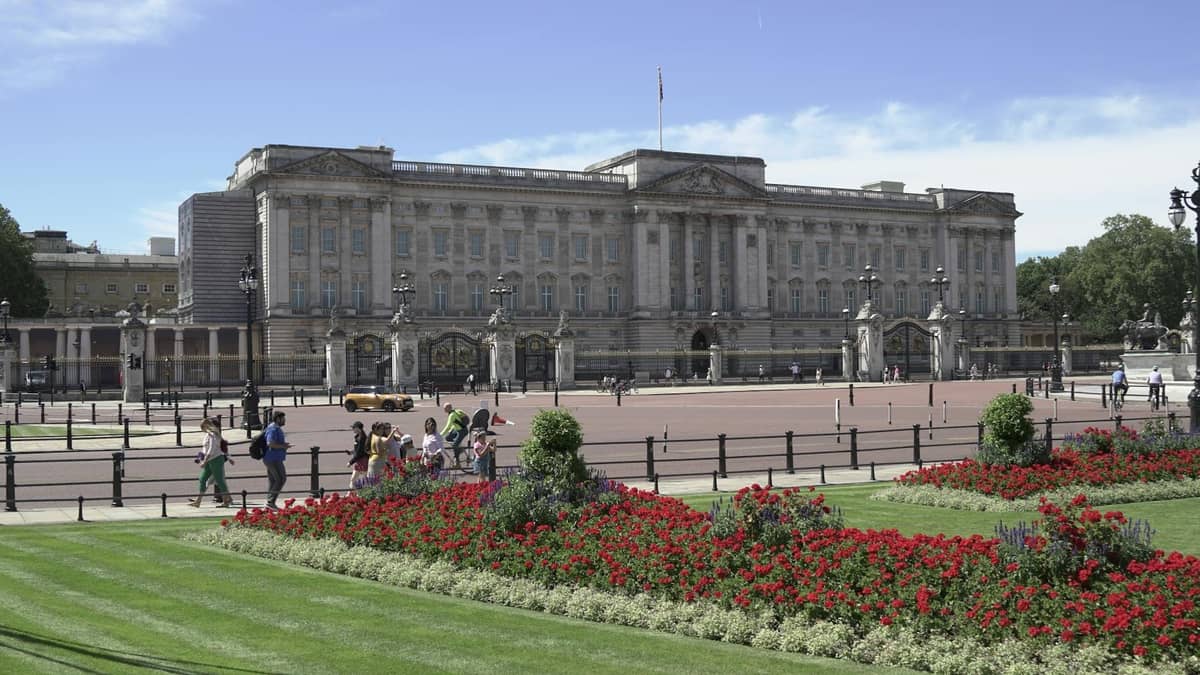 Vista ampla do Buckingham Palace com sua imponente fachada, pátio frontal, portões ornamentados e canteiros de flores coloridos em um dia ensolarado.