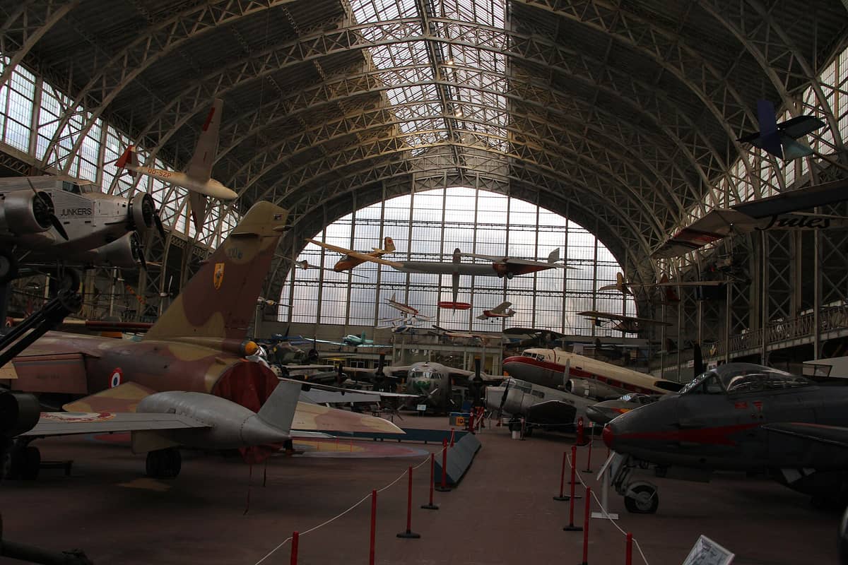 Interior do Museu Real das Forças Armadas e da História Militar em Bruxelas, com aeronaves militares históricas e grandes janelas em arco no hangar de aviação.