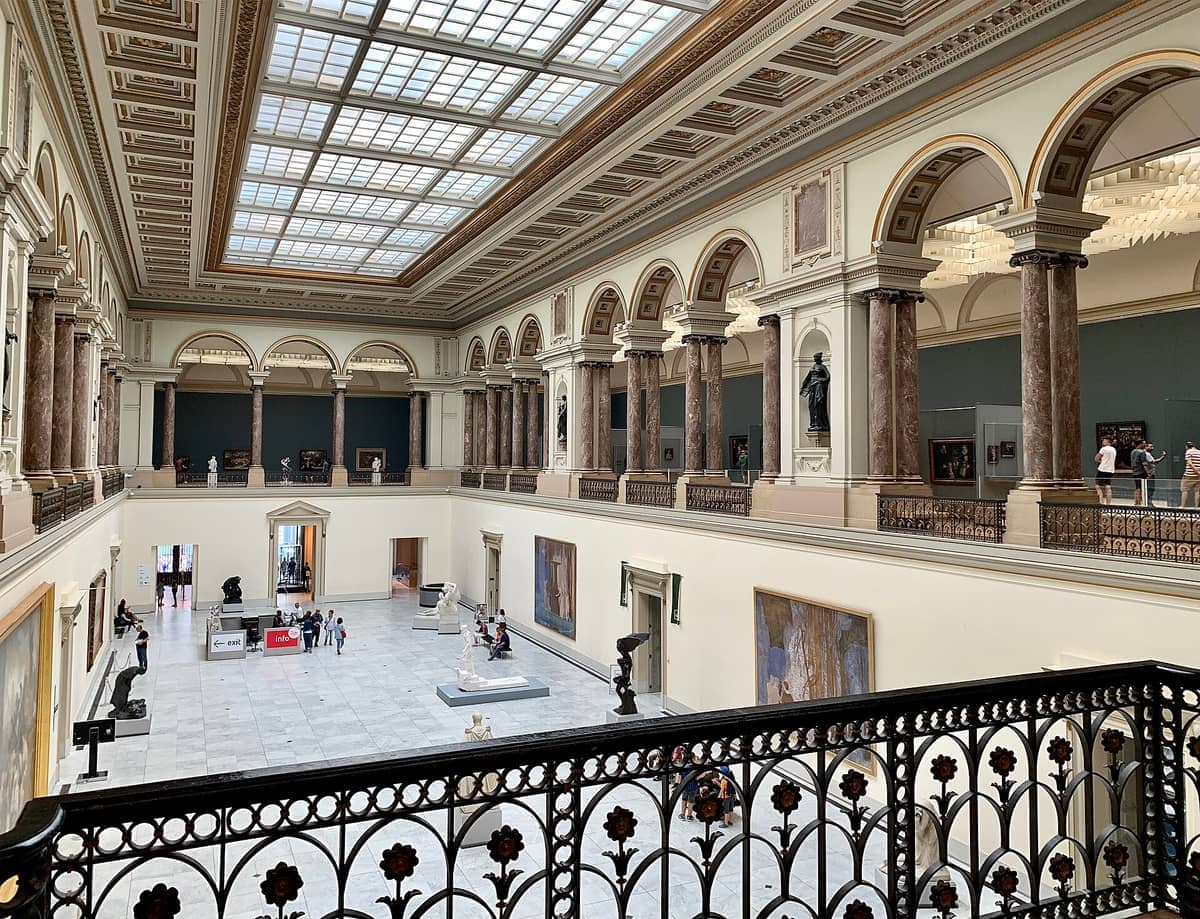 Grande salão dos Museus Reais de Belas-Artes da Bélgica com colunas de mármore, tetos abobadados e visitantes explorando esculturas e pinturas.