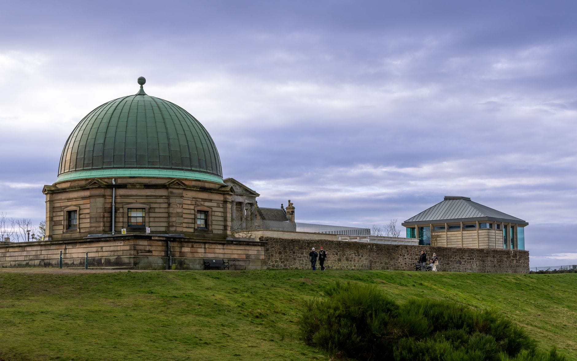Das Royal Observatory Edinburgh mit seiner grünen Kupferkuppel steht auf dem Blackford Hill – im Vordergrund Grashänge und Spaziergänger unter einem bewölkten Himmel.