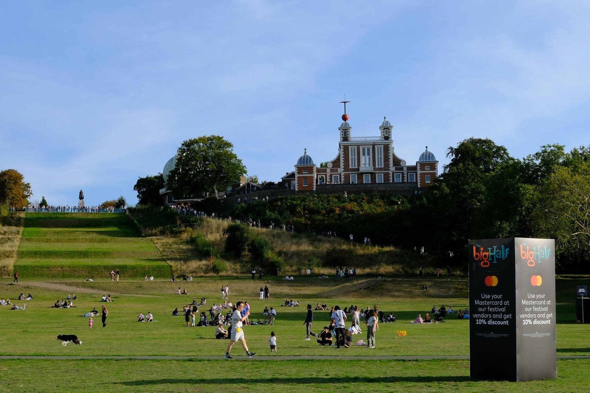 Persone che si rilassano sul prato ai piedi del Royal Observatory Greenwich, con la rossa Time Ball ben visibile contro un cielo azzurro e gli alberi verdi tutt'intorno.