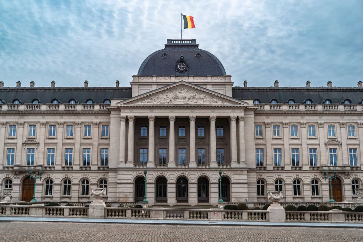 Vista frontal do Palácio Real de Bruxelas com a bandeira belga, colunas neoclássicas e praça de paralelepípedos vazia sob um céu azul com nuvens.