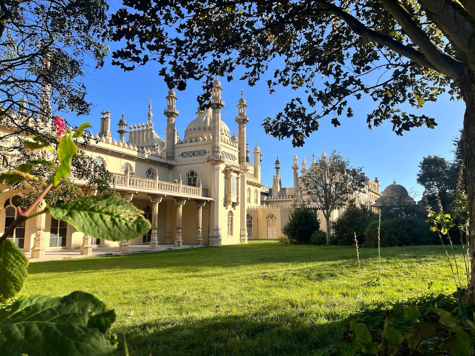 The Royal Pavilion in Brighton seen through leafy trees, framed by lush green Regency-style gardens and a bright blue sky.