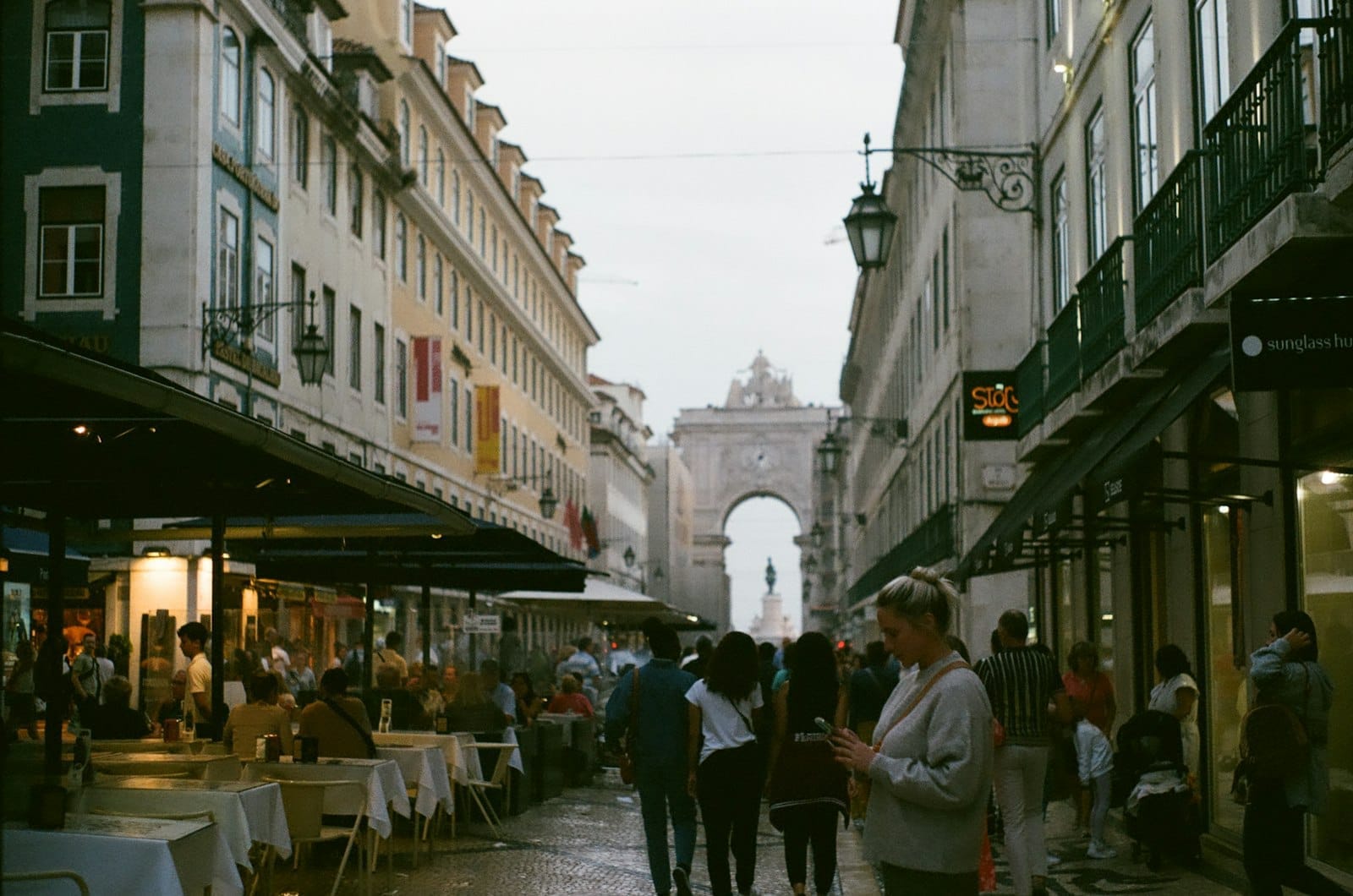Passanti che camminano lungo la Rua Augusta, una strada acciottolata fiancheggiata da caffè e negozi, con l'arco trionfale sullo sfondo.