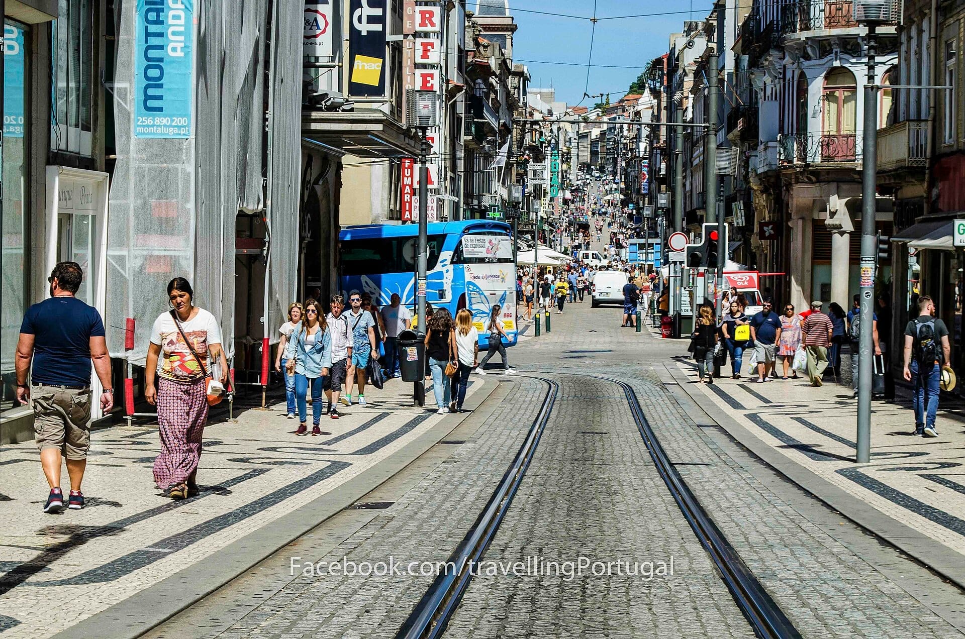 Rua de Santa Catarina a Porto in una giornata di sole, con pedoni che camminano lungo negozi, caffè e binari del tram fiancheggiati da edifici storici.