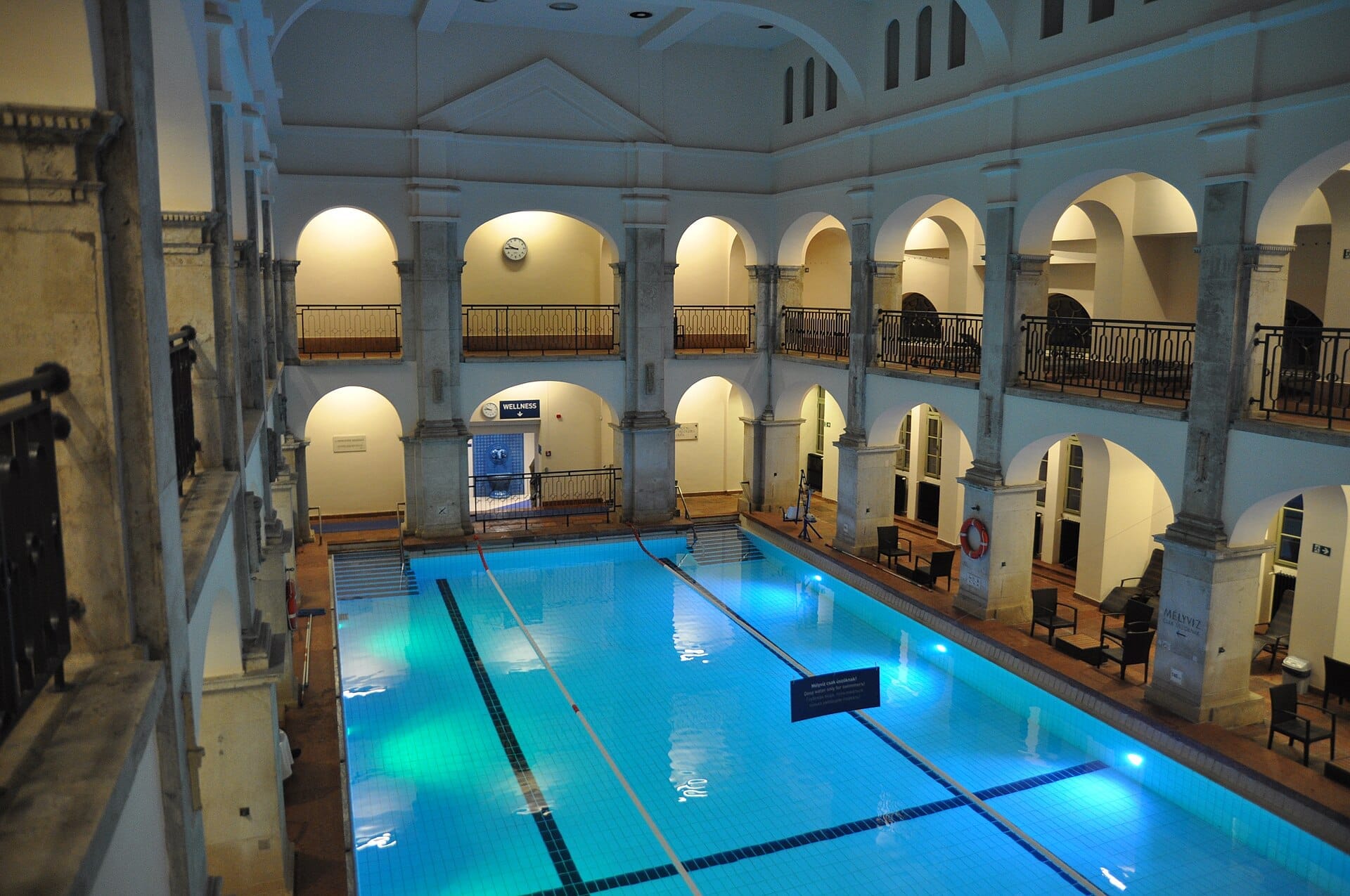 Piscina coperta alle Terme Rudas a Budapest, circondata da balconi ad arco, colonne di pietra e acqua blu con una luce soffusa.