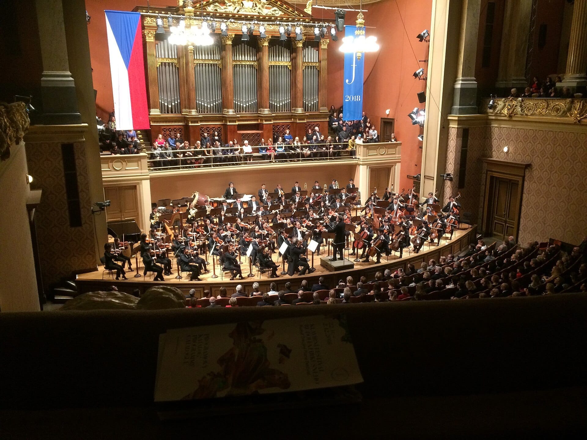Orchestra completa in esibizione sul palco del Rudolfinum, Praga, con la bandiera ceca, canne d'organo dorate e una sala da concerto gremita di pubblico.