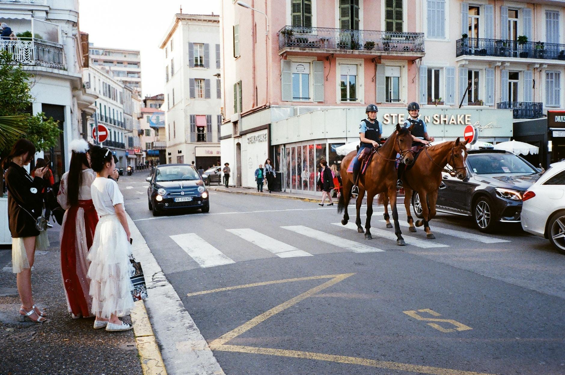 Rue d'Antibes a Cannes con edifici dai colori pastello, persone in costume, auto di passaggio e polizia a cavallo che pattuglia una vivace scena di strada.