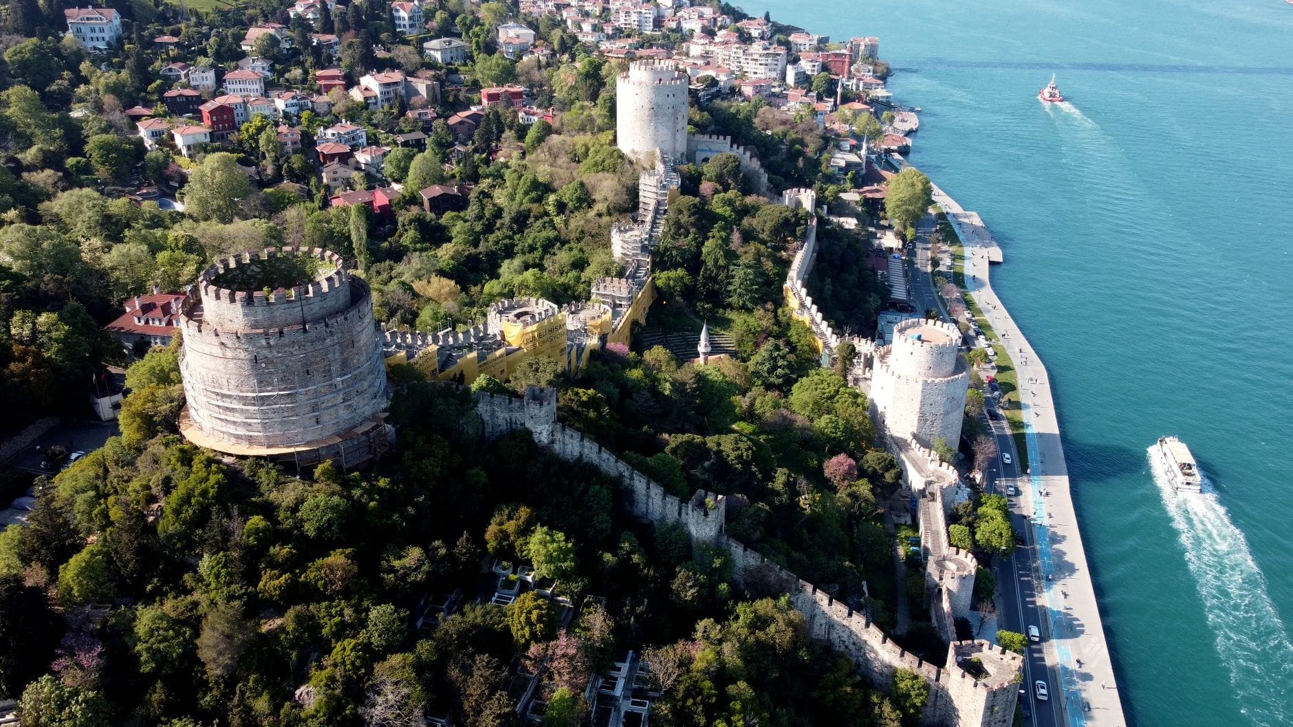 Veduta aerea spettacolare della Fortezza di Rumeli con le sue imponenti torri, le mura storiche, la rigogliosa vegetazione e il Bosforo adiacente, con imbarcazioni e palazzi della città sullo sfondo.
