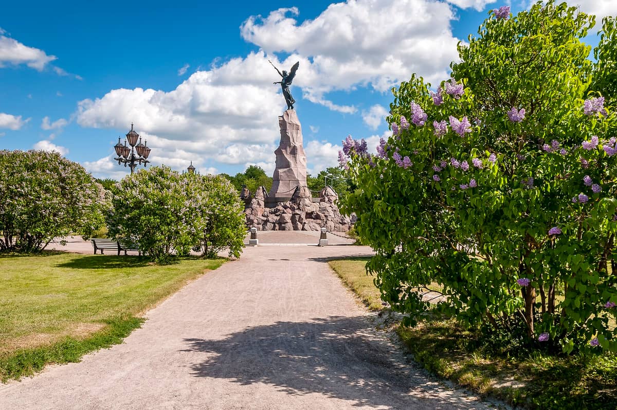 Das Russalka-Denkmal in Tallinn an einem sonnigen Tag – der bronzene Engel auf seiner Granitsäule, umgeben von grünen Bäumen und blühenden Flieder.