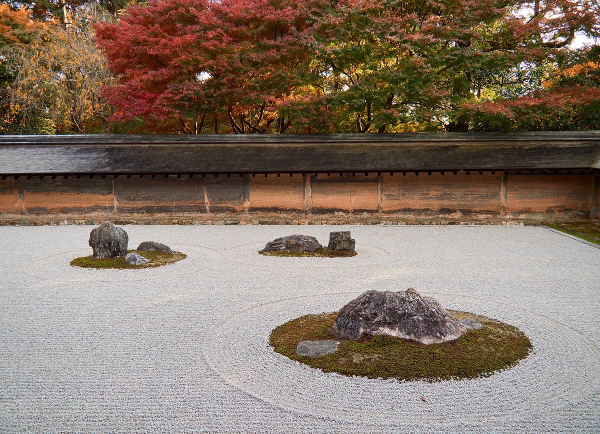 Ryoan-ji’s iconic Zen rock garden with carefully placed stones surrounded by raked white gravel and a backdrop of colorful autumn trees.