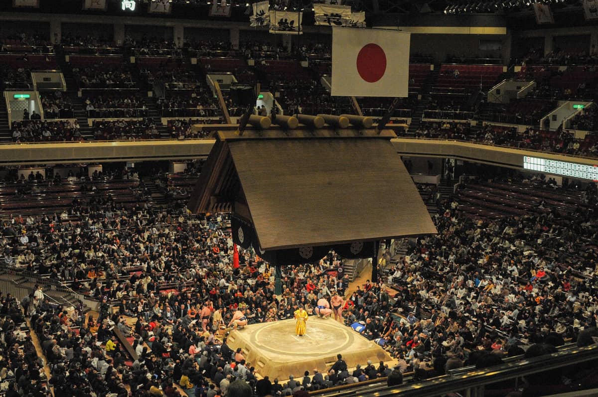 Wide view inside Ryogoku Kokugikan sumo arena with a packed audience watching a sumo match beneath the hanging Japanese flag and traditional roof.