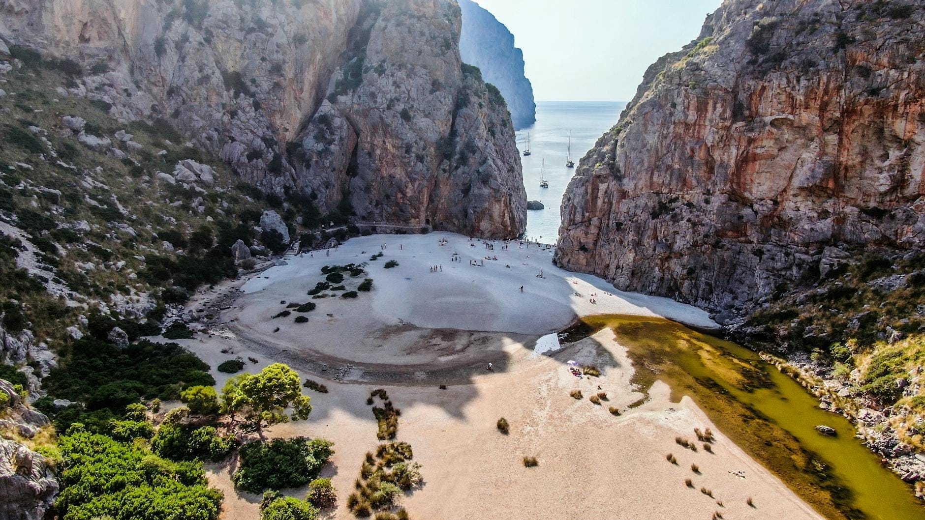 Una spettacolare gola calcarea si apre su una spiaggia di ciottoli con persone che camminano, mare turchese, scogliere a picco e barche ancorate in lontananza a Sa Calobra, Maiorca.