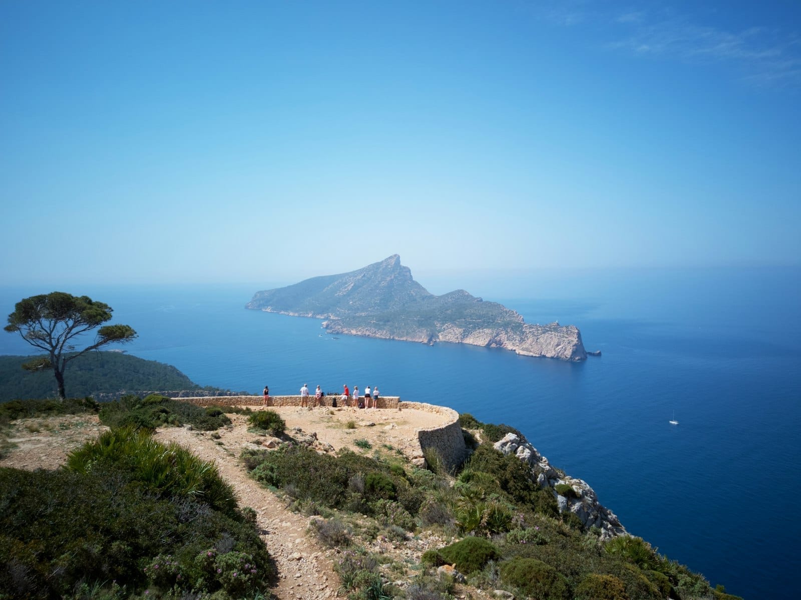 Vista dell'isola Sa Dragonera da un belvedere in cima a una scogliera, con terreno accidentato, mare azzurro e visitatori che godono del paesaggio naturale.