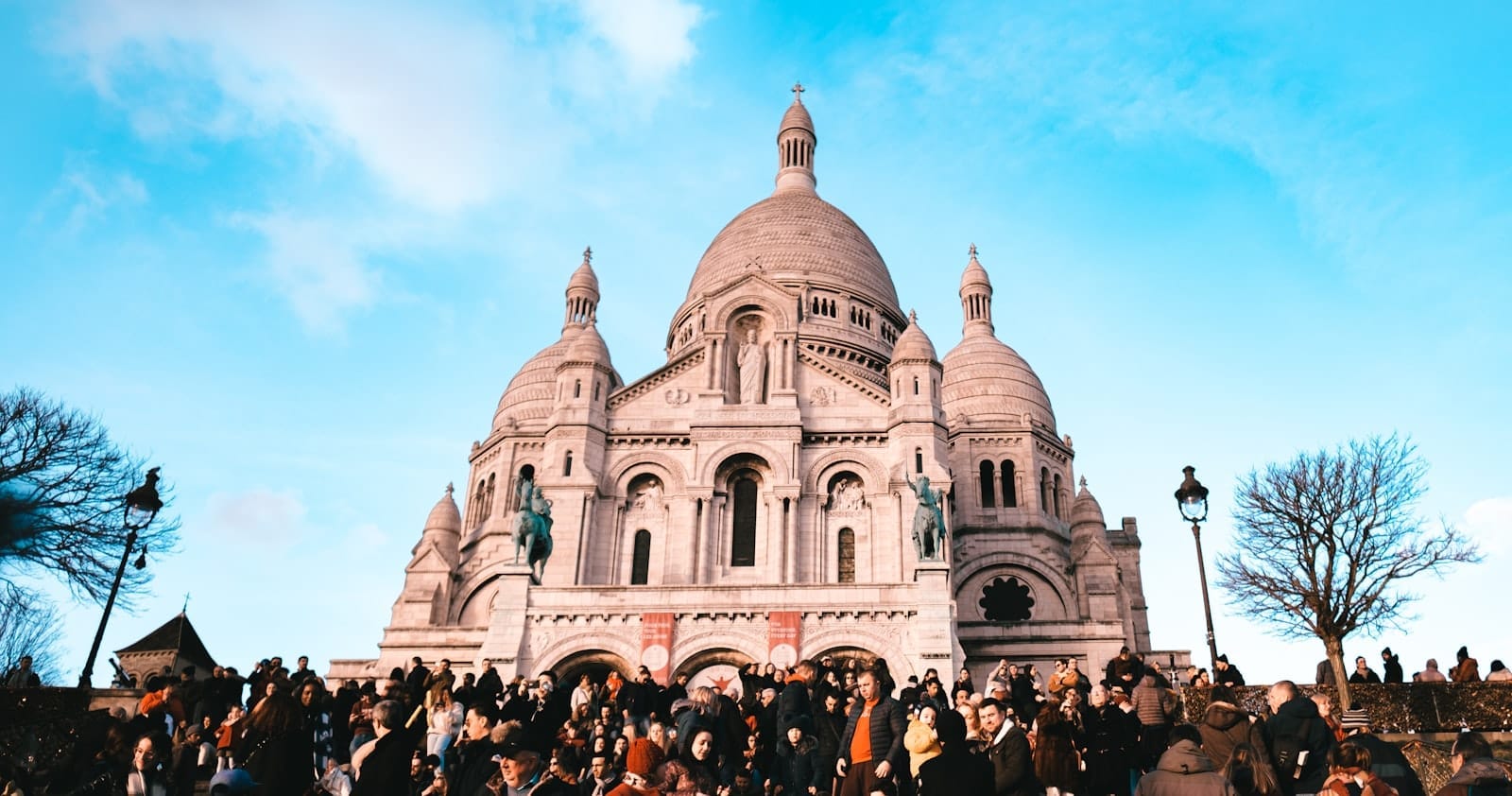 Una folla di visitatori si raduna sui gradini che portano alla Basilica del Sacré-Cœur a Montmartre, sotto un cielo azzurro e luminoso, con le iconiche cupole in bella mostra.