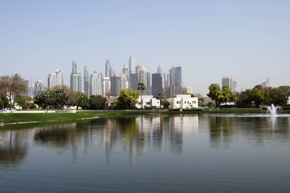 Blick über den Zentralsee im Safa Park mit Dubais Skyline, grünen Rasenflächen, Bäumen und einem Springbrunnen unter klarem Himmel.