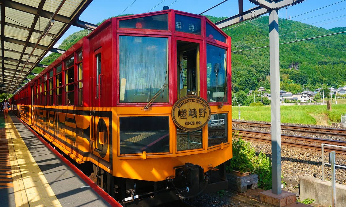 The bright red and yellow Sagano Romantic Train at a countryside station with mountains and lush greenery in the background.