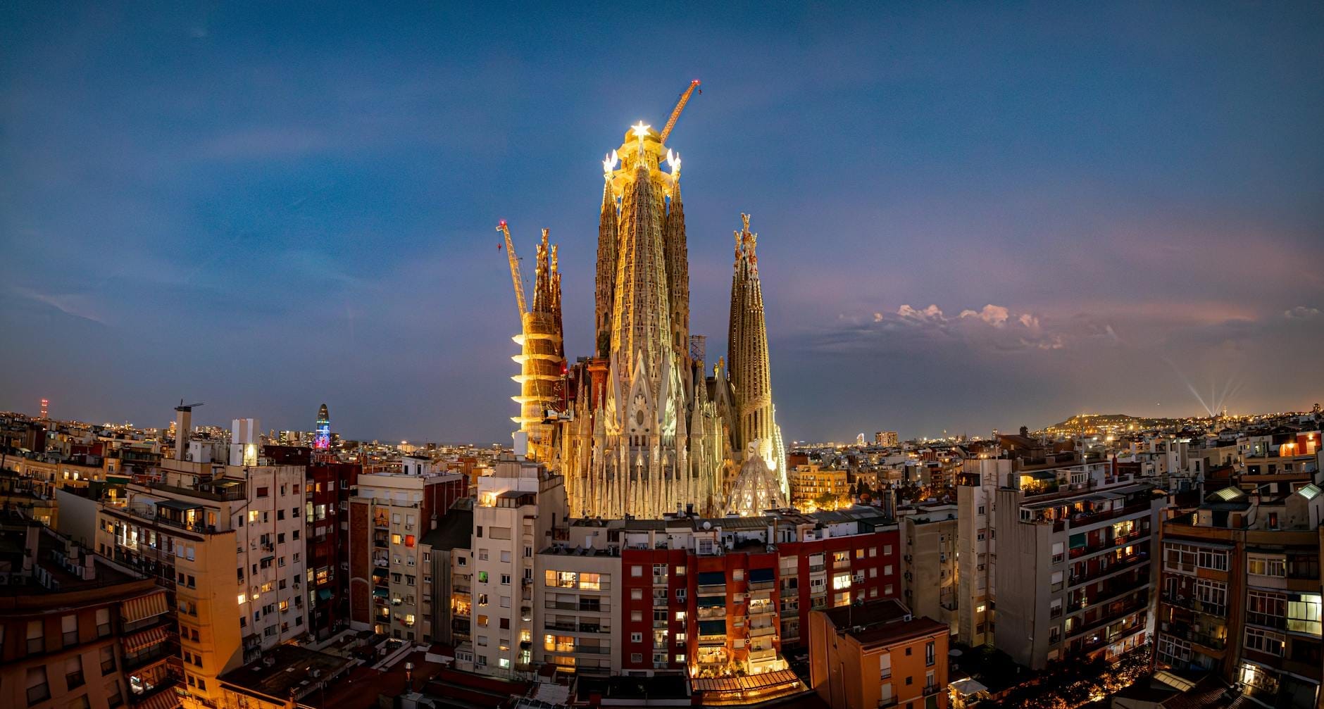 Panoramic evening view of the illuminated Sagrada Família basilica rising above the cityscape of Barcelona, with construction cranes highlighting its ongoing completion.