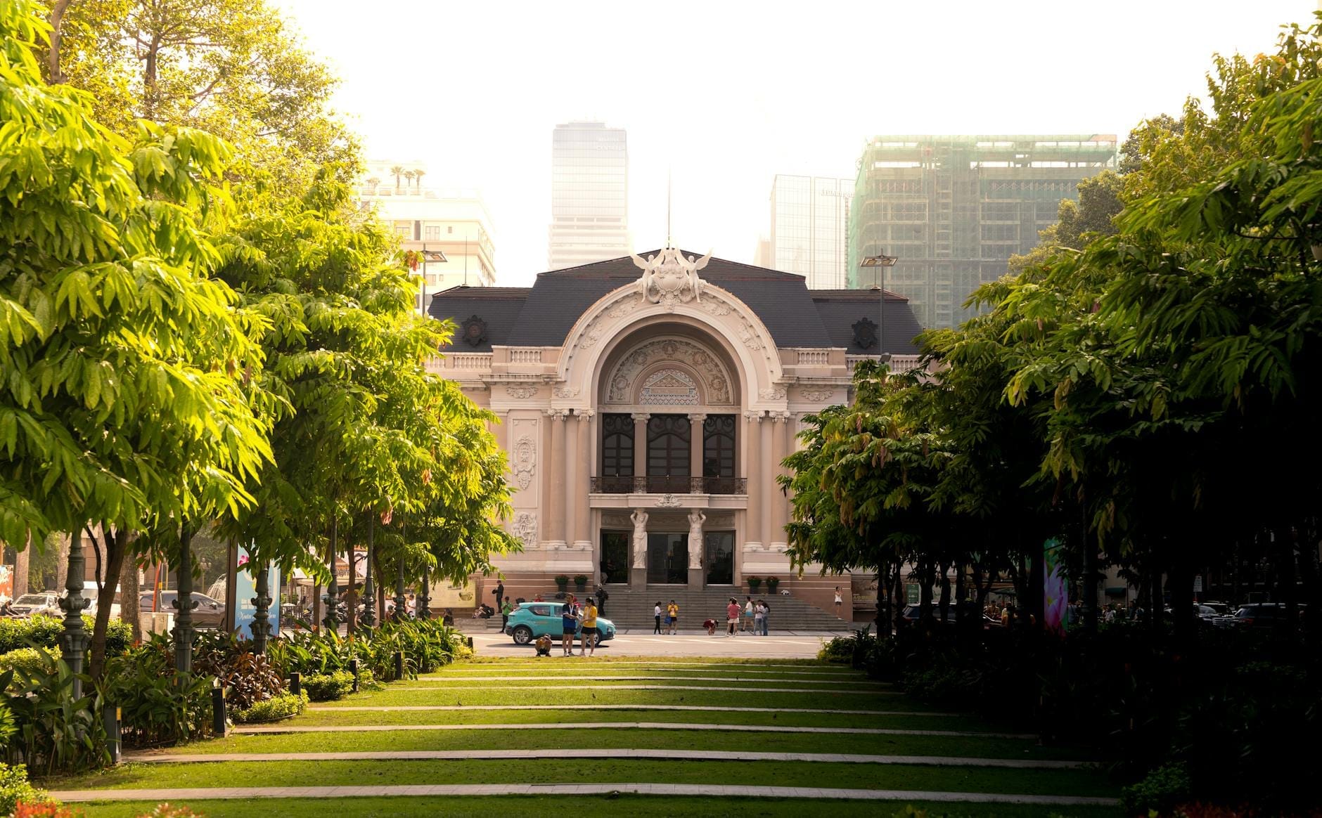 La imponente Ópera de Saigón enmarcada por frondosos árboles verdes y un sendero iluminado por el sol, con edificios modernos de fondo, en una escena vibrante del centro de Ho Chi Minh.
