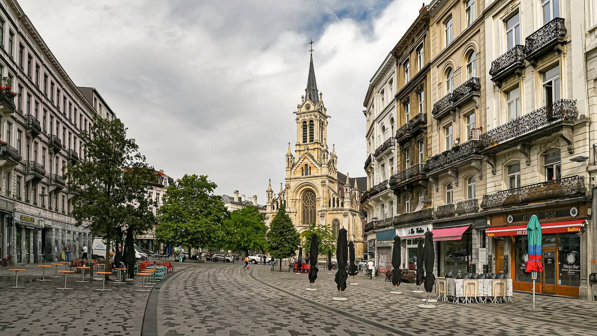 Vista ampla do Parvis de Saint-Gilles em Bruxelas, com suas animadas varandas de café, prédios residenciais e a Igreja de Saint-Gilles ao centro da praça.