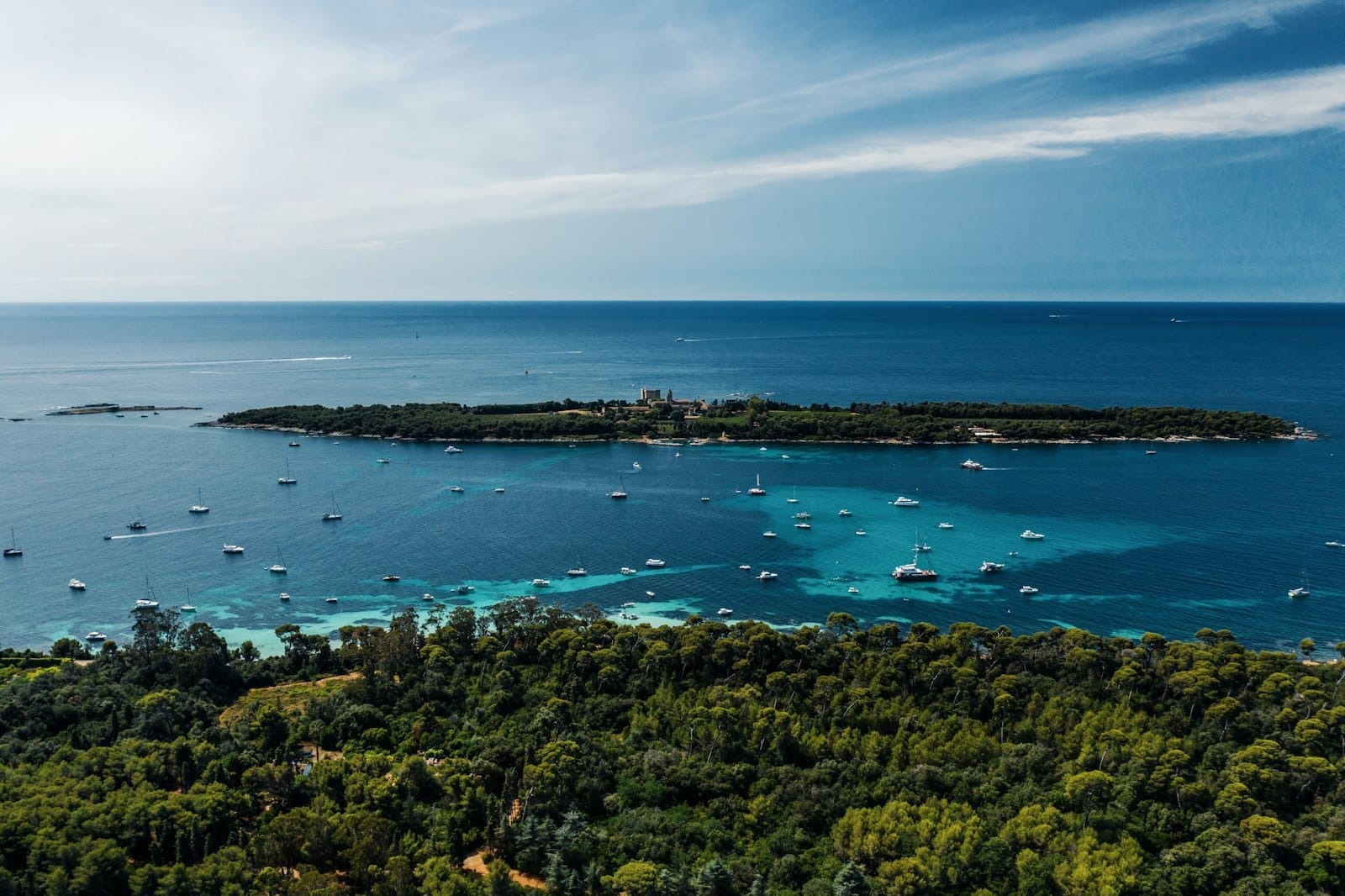 Vista aerea dell'Île Saint-Honorat circondata da acque turchesi e barche, con una lussureggiante vegetazione e il cielo azzurro sopra.