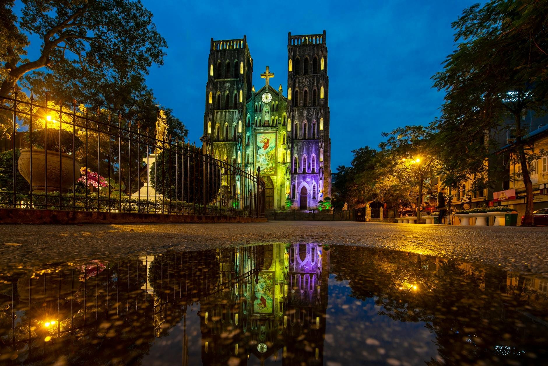 Saint Joseph's Cathedral in Hanoi illuminated at twilight with its twin towers and gothic facade reflected in a puddle, framed by trees and a lively square.
