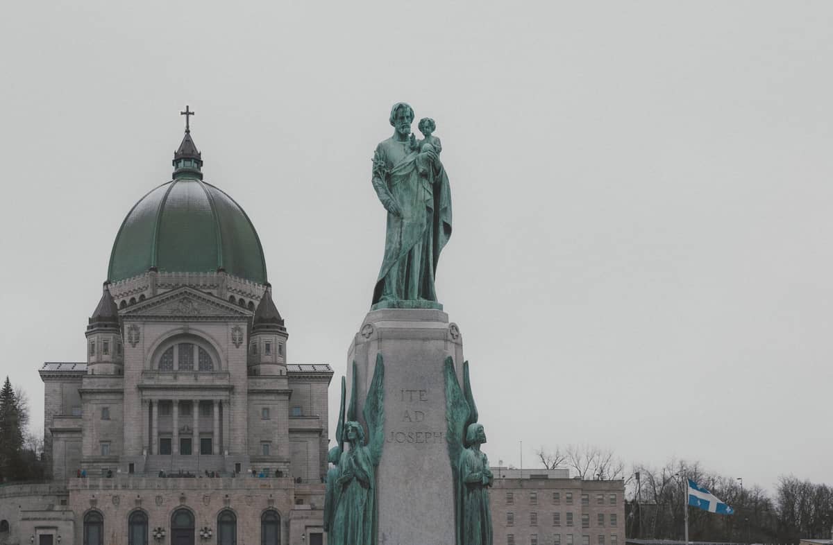 Außenansicht des Oratoire Saint-Joseph du Mont-Royal mit seiner markanten grünen Kupferkuppel und einer Statue des heiligen Josef im Vordergrund an einem bewölkten Tag.
