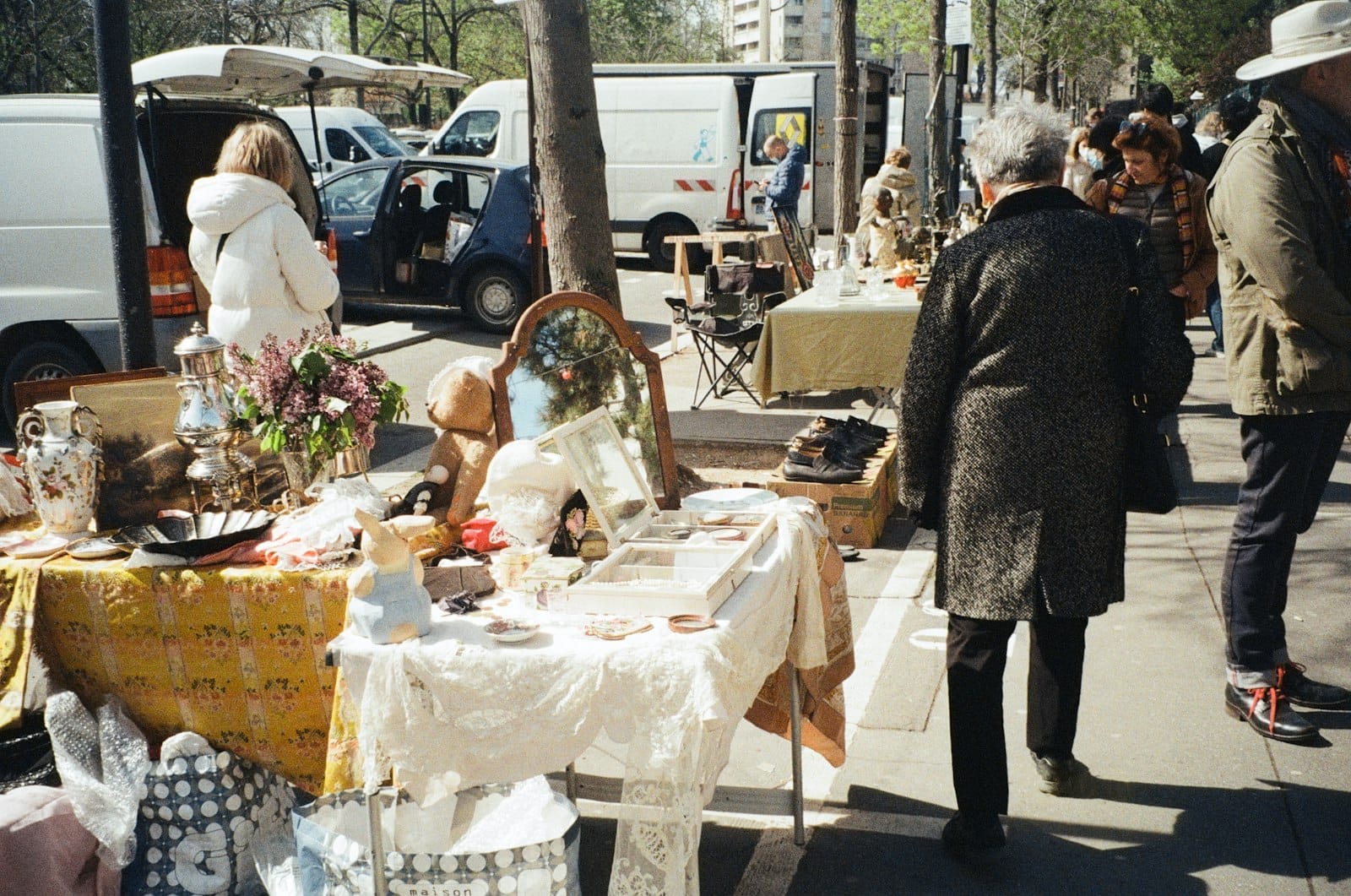 Scena di mercato delle pulci all'aperto con persone che curiosano tra tavoli pieni di oggetti d'antiquariato, pezzi vintage e da collezione lungo una strada parigina in una giornata di sole.