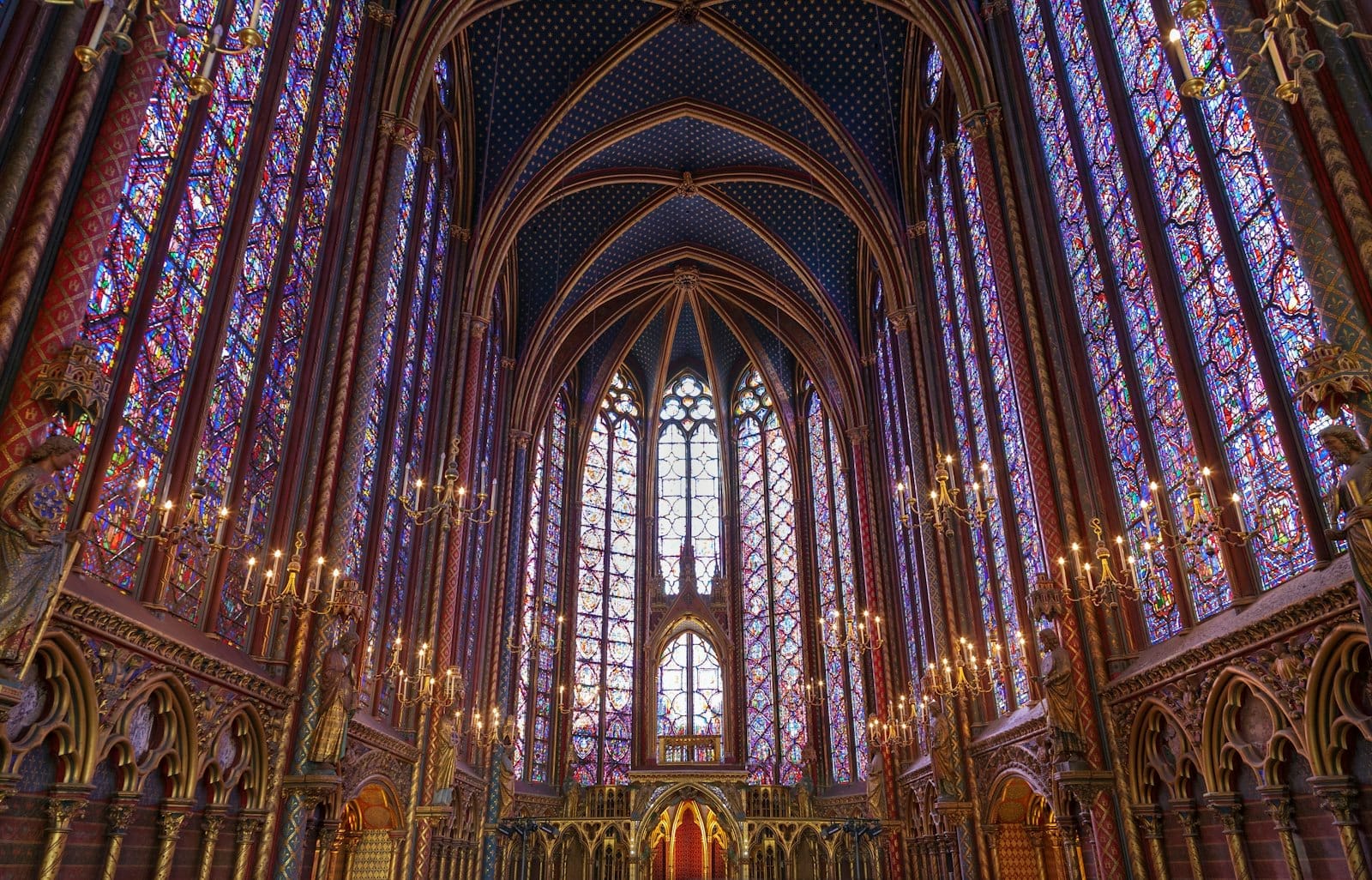 Interno della cappella superiore della Sainte-Chapelle: altissime pareti di vetrate colorate, volte affrescate, lampadari dorati e raffinati elementi gotici in pietra immersi in una luce multicolore.