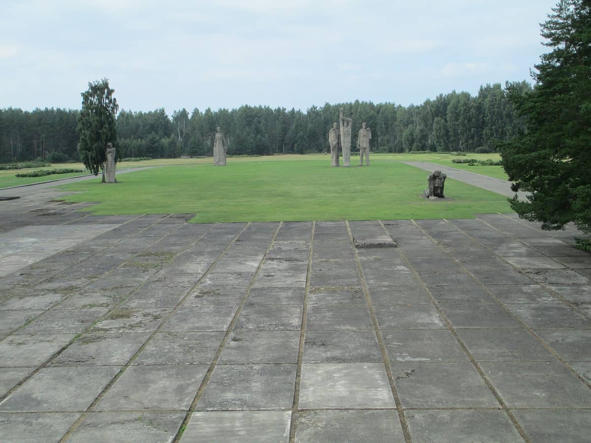 Blick auf die Gedenkstätte Salaspils mit monumentalen Betonskulpturen, weitläufigen grünen Rasenflächen und umgebendem Kiefernwald unter bewölktem Himmel.