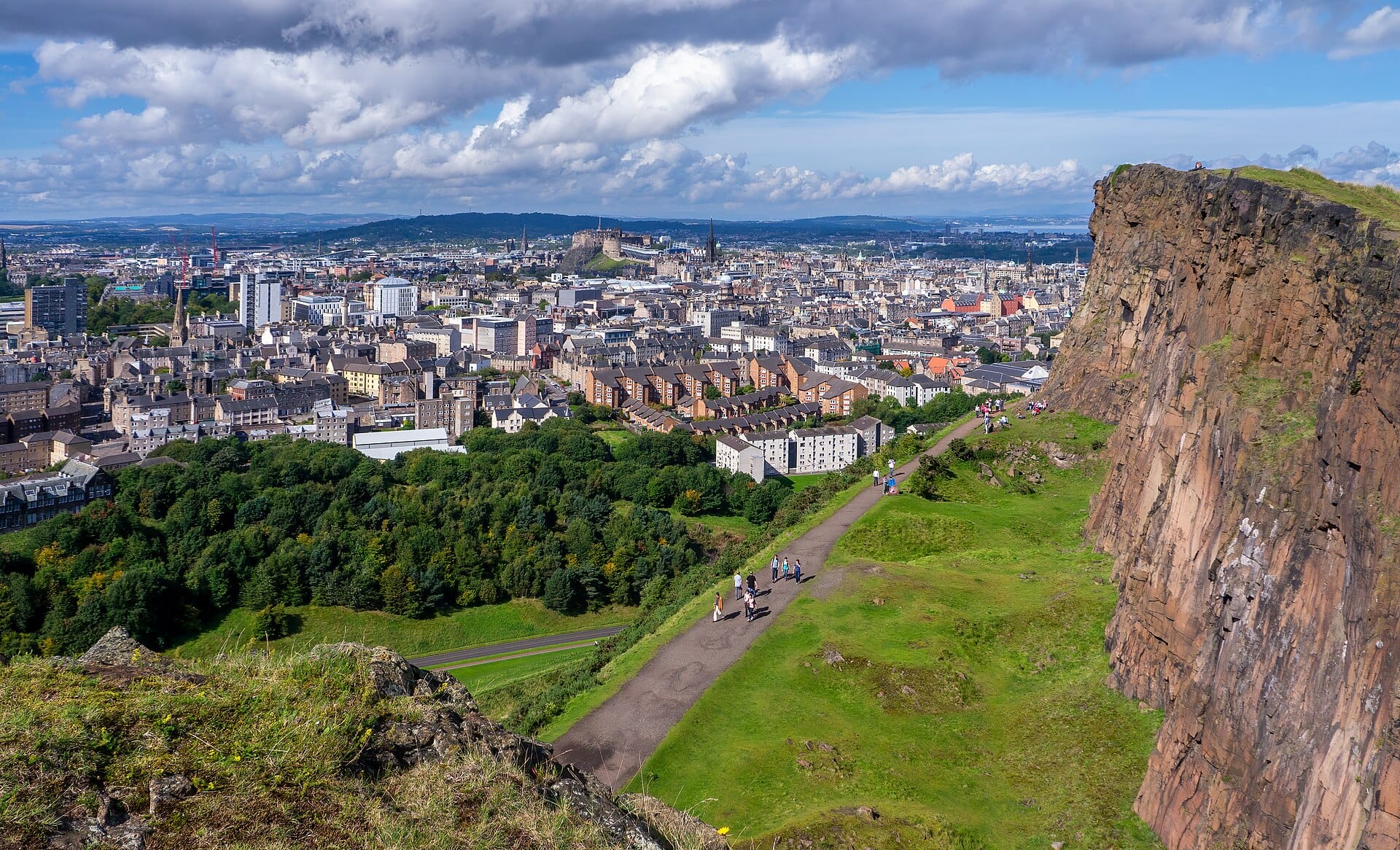 Blick auf die senkrechten Felswände der Salisbury Crags über Edinburgh, mit der Stadt und dem Schloss im Hintergrund bei leicht bewölktem Himmel.