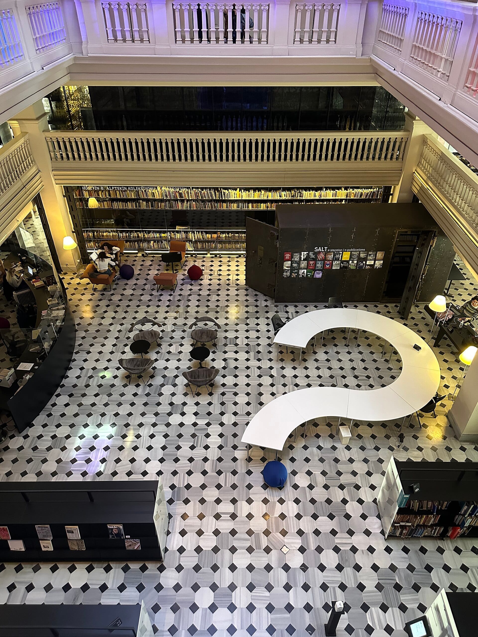 Vista degli interni ariosi di SALT Galata, con pavimento a mosaico, tavoli curvi moderni, librerie e posti a sedere accoglienti sotto un grande mezzanino.