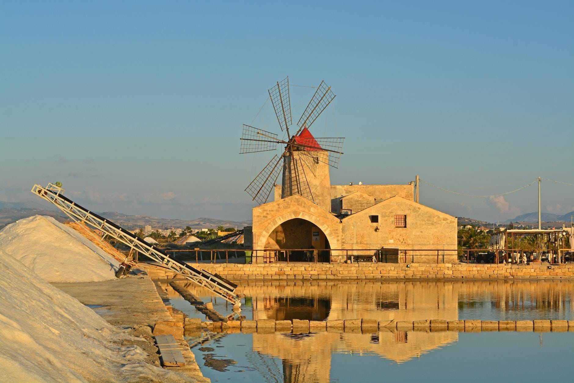 Traditionelle Windmühle und historische Salzwerksgebäude, die sich im Wasser der Salzgärten von Trapani, Sizilien, unter einem klaren Abendhimmel spiegeln.