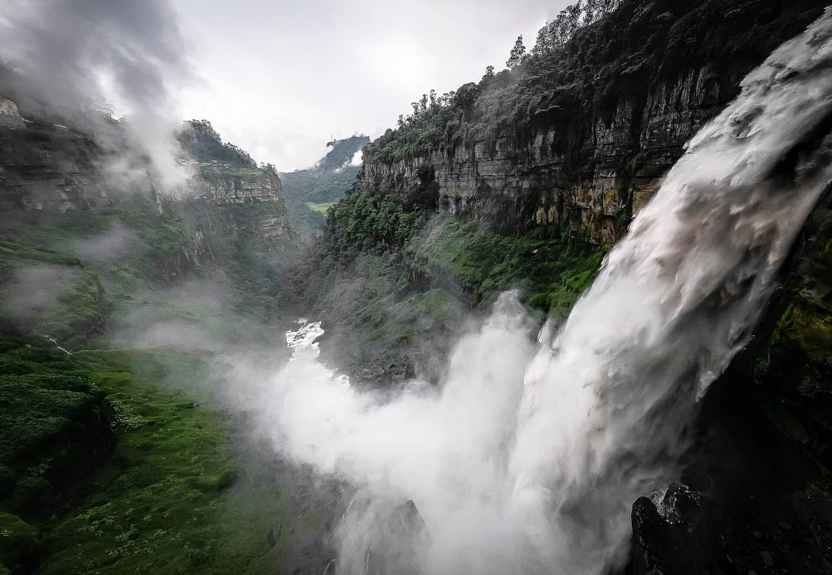Dramatic view of Salto del Tequendama waterfall plunging off a misty cliff into the gorge below, surrounded by lush green forest and rocky canyon walls.