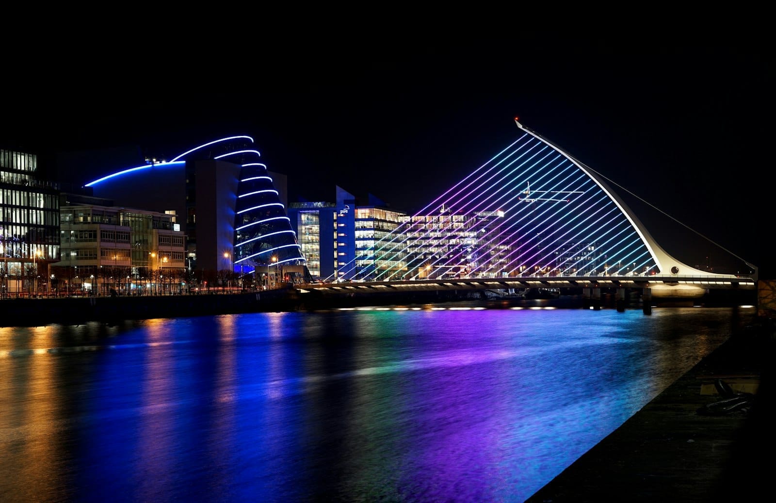 Il Ponte Samuel Beckett illuminato di notte, con i riflessi colorati sul fiume Liffey e gli edifici moderni sullo sfondo.