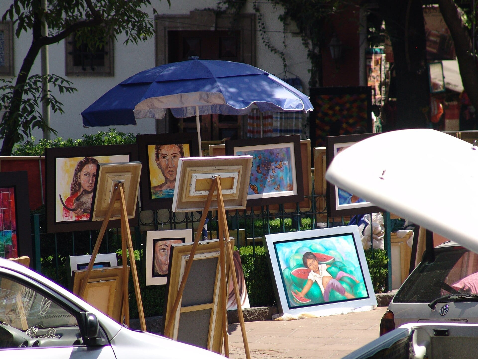 Mercato d'arte all'aperto al Bazar del Sábado a San Ángel, dipinti colorati su cavalletti sotto un ombrellone blu, circondati da auto e vegetazione.